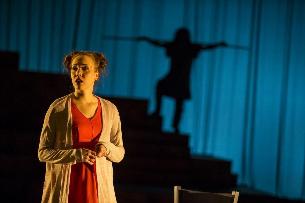 Women stands on stage with a shadow of a man behind her.